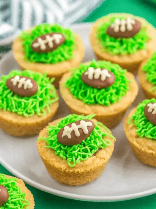 Football-shaped cookie cups filled with green frosting “grass” and topped with chocolate football candies on a white plate, perfect Super Bowl party food for a football-shaped dessert table.