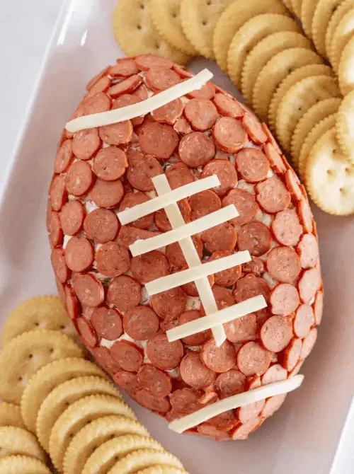 Football-shaped pepperoni pizza cheese ball decorated with mozzarella laces and surrounded by crackers on a white platter, a fun Super Bowl party food centerpiece.