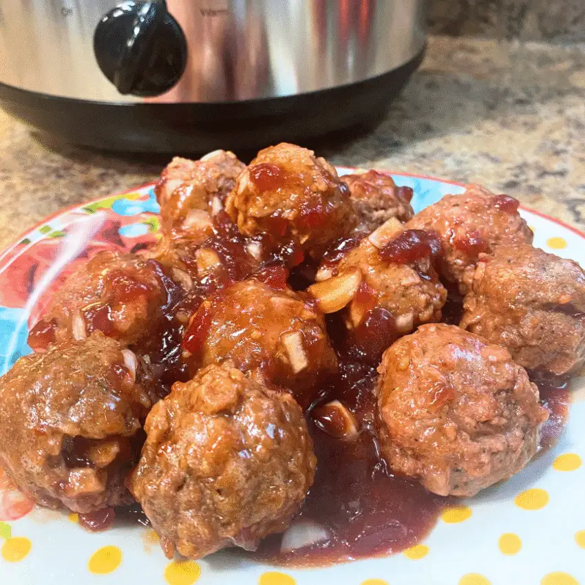 Close-up of sweet chili cranberry crockpot meatballs on a colorful plate, coated in a tangy cranberry and sweet chili sauce, with a slow cooker in the background as an easy holiday appetizer for a crowd.