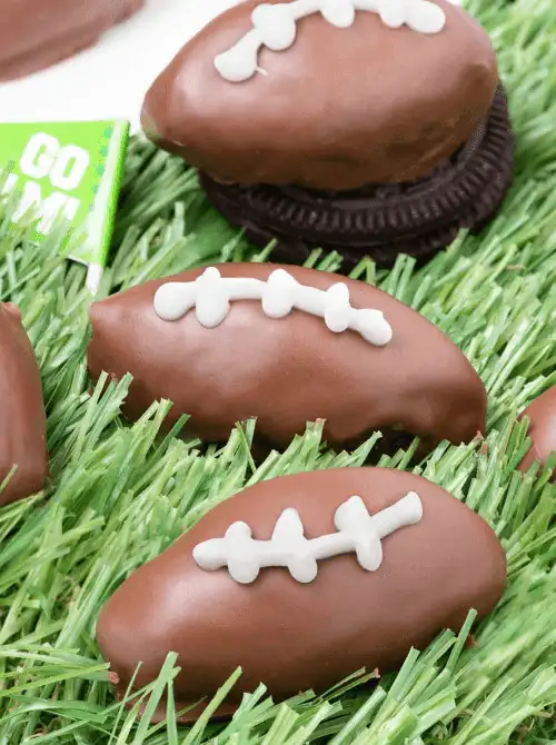 Football-shaped Oreo balls coated in chocolate and decorated with white icing laces displayed on faux grass, perfect Super Bowl party food for a football-shaped dessert display.