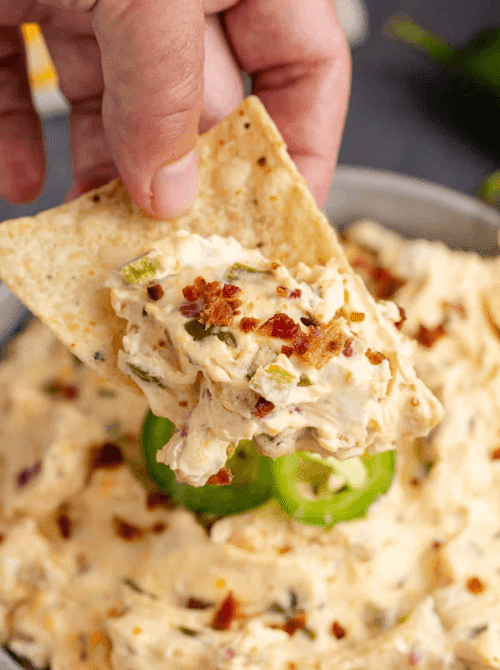 A tortilla chip dipping into creamy jalapeño chicken dip with bacon and sliced peppers, perfect for easy crock pot recipes at a last minute Super Bowl party.
