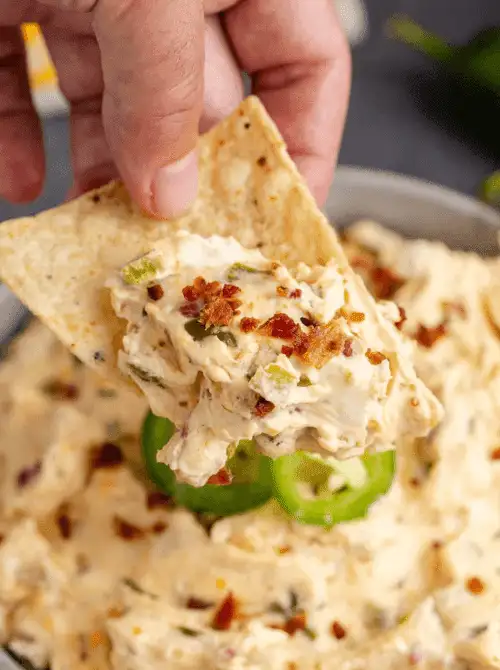 A tortilla chip dipping into creamy jalapeño chicken dip with bacon and sliced peppers, perfect for easy crock pot recipes at a last minute Super Bowl party.