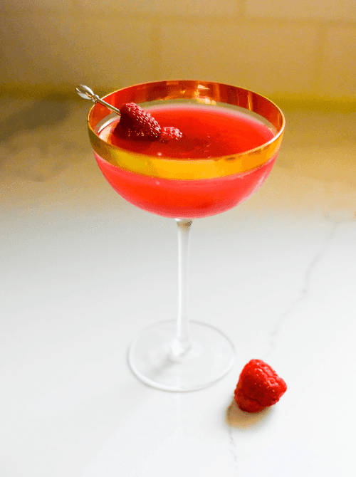 Pink Cocktail Ideas for a Galentine’s Day Party featuring a bright raspberry martini in a coupe glass with gold rim and fresh raspberry garnish