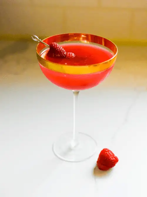 Pink Cocktail Ideas for a Galentine’s Day Party featuring a bright raspberry martini in a coupe glass with gold rim and fresh raspberry garnish