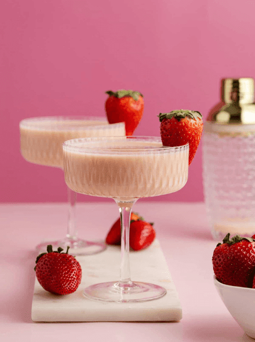 Pink cocktail ideas for a Galentine’s Day party featuring creamy strawberry martinis garnished with fresh strawberries on a pink background.