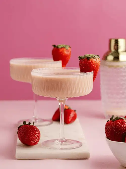 Pink cocktail ideas for a Galentine’s Day party featuring creamy strawberry martinis garnished with fresh strawberries on a pink background.