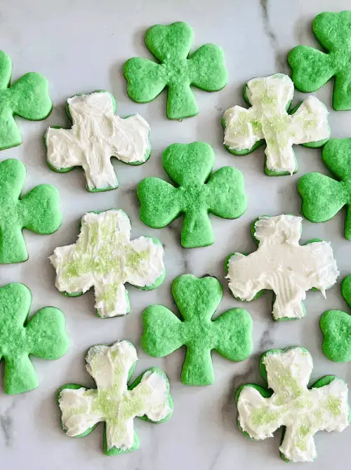 Overhead view of green shamrock sugar cookies, some frosted with white icing and sparkling sugar, arranged for festive St. Patrick’s Day Dessert Ideas.