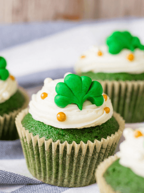 Green velvet cupcakes with creamy white frosting and shamrock toppers arranged for festive St. Patrick’s Day Dessert Ideas, accented with gold sugar pearls.