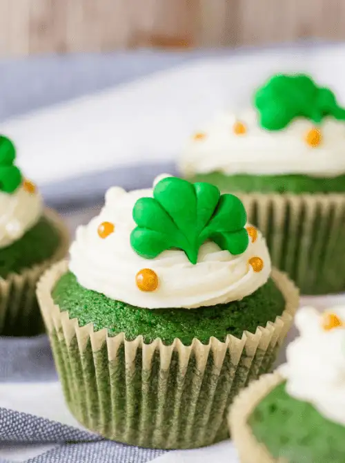 Green velvet cupcakes with creamy white frosting and shamrock toppers arranged for festive St. Patrick’s Day Dessert Ideas, accented with gold sugar pearls.