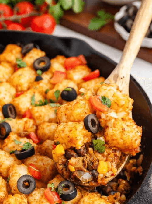Mexican-inspired tater tot casserole in a cast iron skillet topped with black olives, diced tomatoes, and cilantro, a flavorful example of super easy casseroles and creative family dinner ideas.