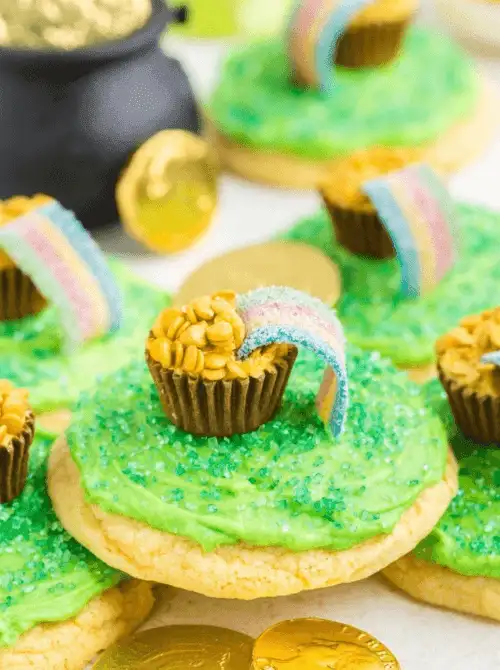 Green frosted sugar cookies decorated with rainbow candy arches, mini peanut butter cups, and gold coins styled for playful St. Patrick’s Day Dessert Ideas at a themed party table. 🍀
