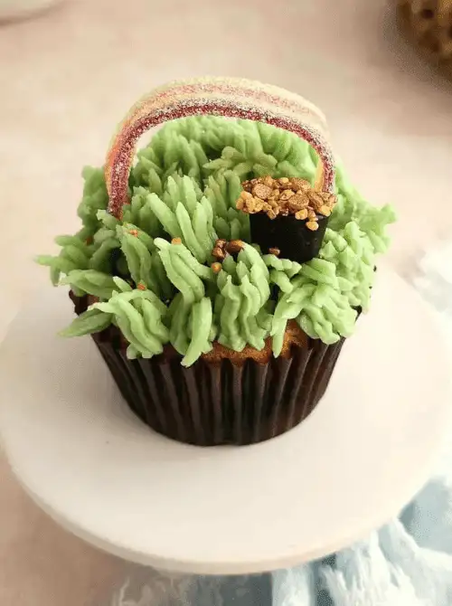 Shamrock-inspired cupcake with green grass-style frosting, rainbow candy strip, and gold sprinkle accents displayed as part of creative St. Patrick’s Day Dessert Ideas.
