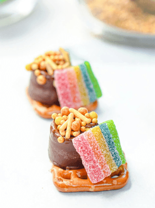 Mini Rolo pretzel bites topped with rainbow candy strips and gold sprinkles arranged in a row for playful St. Patrick’s Day Dessert Ideas on a snack table. 🍀