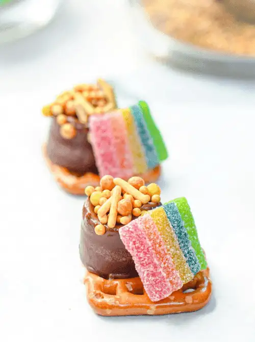Mini Rolo pretzel bites topped with rainbow candy strips and gold sprinkles arranged in a row for playful St. Patrick’s Day Dessert Ideas on a snack table. 🍀