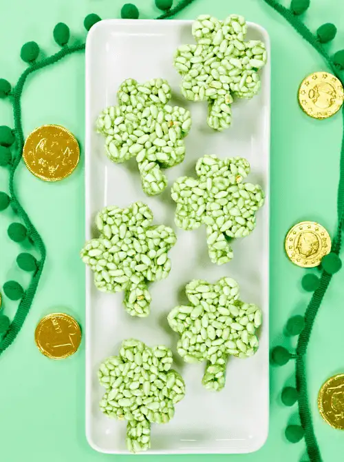 Shamrock-shaped green Rice Krispie treats arranged on a white platter with gold coins, featured in festive St. Patrick’s Day Dessert Ideas for a themed party table. 🍀