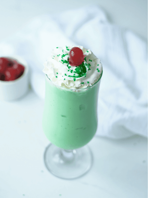 Mint green shamrock shake topped with whipped cream, green sprinkles, and a cherry in a tall glass, an easy no-bake St. Patrick’s Day dessert on a budget.