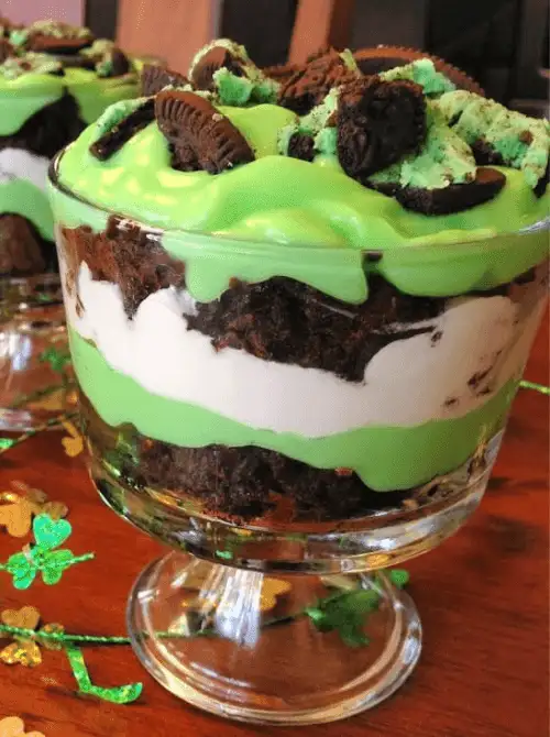 Layered brownie trifle with green pudding, whipped cream, and Oreo pieces in a glass dish, a make-ahead dessert for St. Patrick’s Day and desserts on a budget.