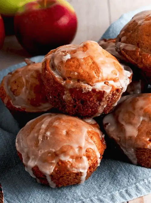 Glazed apple breakfast muffins stacked on a plate with fresh apples in the background, a sweet bakery-style treat to serve alongside make-ahead Easter brunch casseroles.