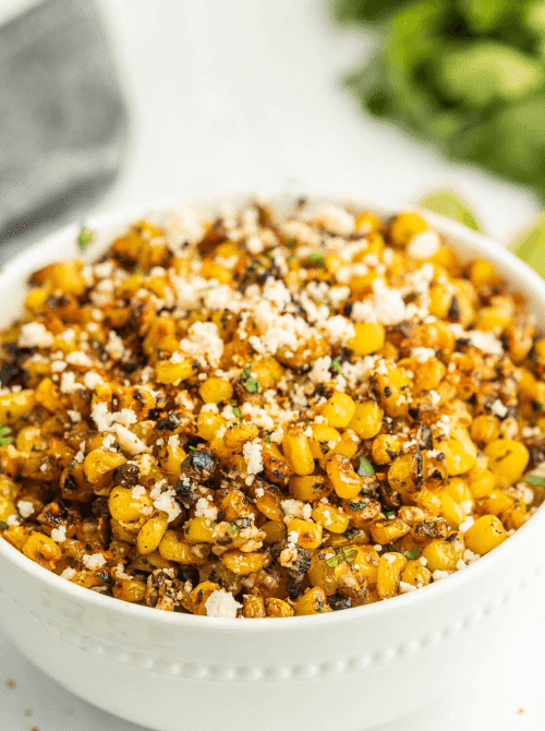Blackstone Mexican street corn off the cob in a white bowl with charred kernels, cotija cheese, and cilantro — an easy Cinco de Mayo party food recipe for a crowd that's ready in just 20 minutes.