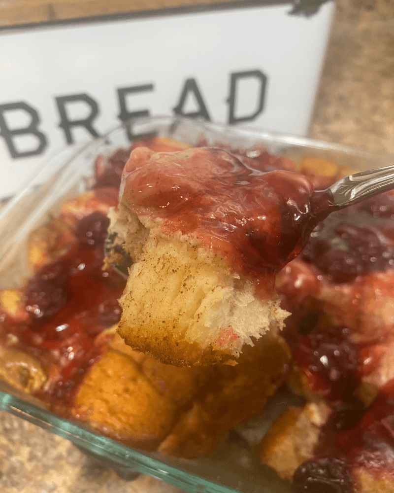 Close-up of a spoon lifting a warm square of Cherry Cheesecake Monkey Bread Casserole from a glass baking dish, showing soft cinnamon biscuit layers topped with glossy cherry pie filling.
