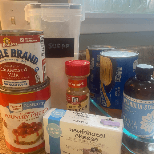 Ingredients for Cherry Cheesecake Monkey Bread Casserole including refrigerated biscuit dough, cherry pie filling, sweetened condensed milk, sugar, ground cinnamon, vanilla extract, and Neufchâtel cream cheese arranged on a kitchen counter.