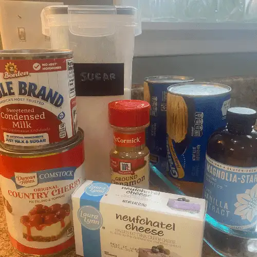 Ingredients for Cherry Cheesecake Monkey Bread Casserole including refrigerated biscuit dough, cherry pie filling, sweetened condensed milk, sugar, ground cinnamon, vanilla extract, and Neufchâtel cream cheese arranged on a kitchen counter.
