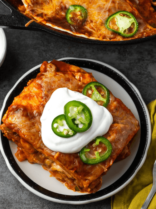 One pan chicken enchilada skillet slice on a white plate topped with sour cream and fresh jalapeño slices with a cast iron skillet in the background — an easy Cinco de Mayo party food recipe for a crowd that feeds 8 from a single pan for just $15.