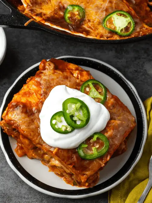 One pan chicken enchilada skillet slice on a white plate topped with sour cream and fresh jalapeño slices with a cast iron skillet in the background — an easy Cinco de Mayo party food recipe for a crowd that feeds 8 from a single pan for just $15.