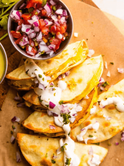 Crispy potato tacos de papa stacked on parchment paper drizzled with creamy jalapeño sauce and diced red onion with a bowl of pico de gallo on the side — a budget-friendly easy Cinco de Mayo party food recipe for a crowd that makes 18 tacos for under $9.