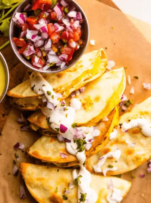 Crispy potato tacos de papa stacked on parchment paper drizzled with creamy jalapeño sauce and diced red onion with a bowl of pico de gallo on the side — a budget-friendly easy Cinco de Mayo party food recipe for a crowd that makes 18 tacos for under $9.