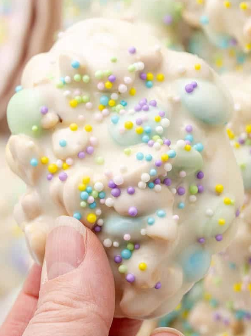 Hand holding a pastel no-bake Easter-themed dessert for a bake sale, featuring white chocolate–coated clusters with colorful Easter candy eggs and festive spring sprinkles.