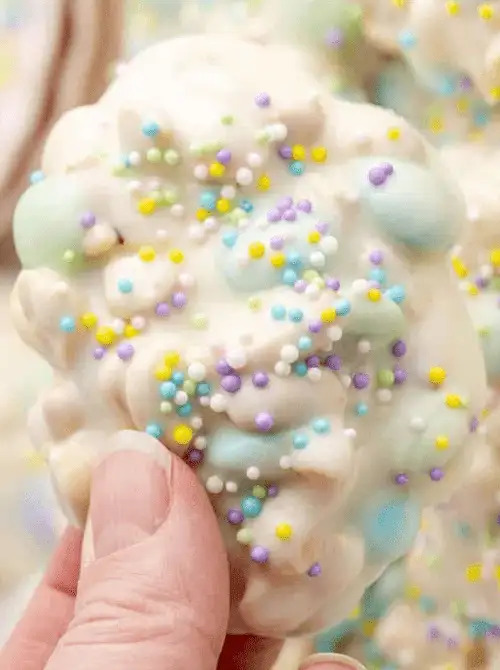 Hand holding a pastel no-bake Easter-themed dessert for a bake sale, featuring white chocolate–coated clusters with colorful Easter candy eggs and festive spring sprinkles.