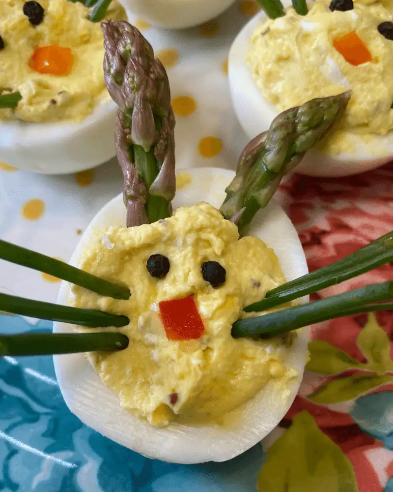 Overhead close-up of a single Easter Bunny Deviled Egg on a yellow polka dot plate with a colorful floral background. The egg is filled with a generous mound of creamy yellow filling and decorated with two asparagus spear ears, long chive whiskers extending to both sides, a red pepper square nose, and two black peppercorn eyes, with additional bunny eggs softly visible in the background.