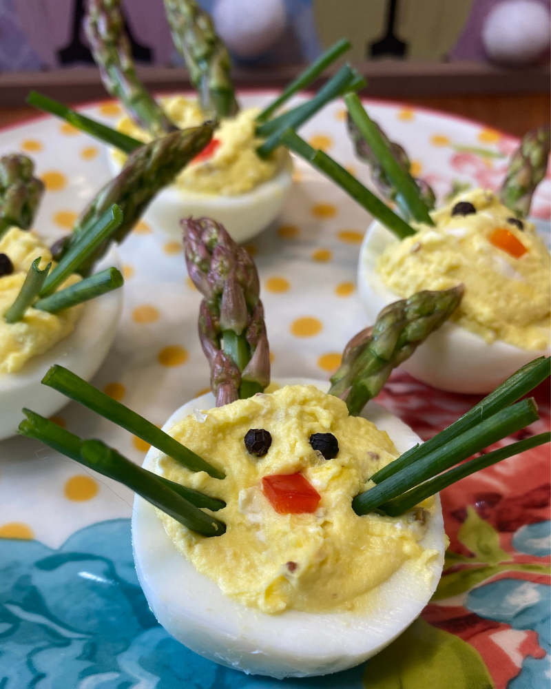 Close-up of Easter Bunny Deviled Eggs on a colorful floral and yellow polka dot plate. The foreground egg shows a creamy yellow filling decorated with asparagus spear ears, green chive whiskers, a red pepper square nose, and two black peppercorn eyes, with several more bunny eggs visible behind it.