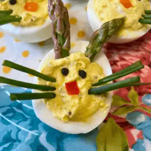Close-up of Easter Bunny Deviled Eggs arranged on a blue and coral spring platter. Each egg half is filled with a creamy yellow filling and decorated with asparagus spear ears, chive whiskers, a red pepper square nose, and black peppercorn eyes.