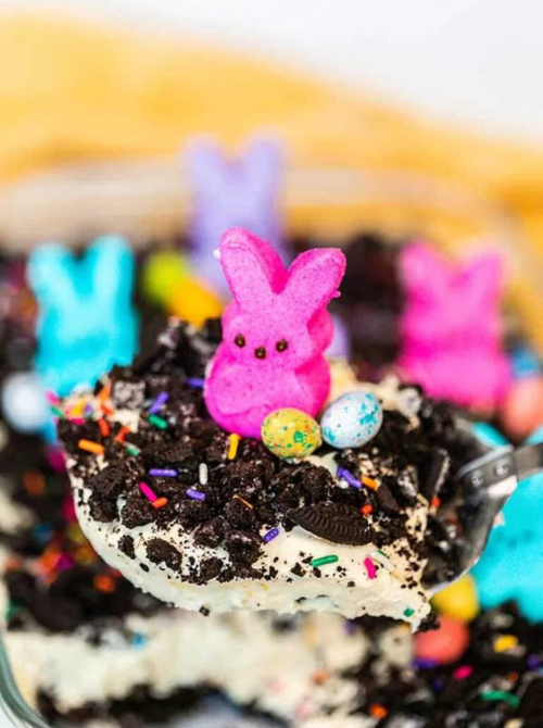 Spoonful of creamy no-bake Easter-themed dessert for a bake sale, topped with crushed chocolate cookies, colorful sprinkles, candy eggs, and a bright marshmallow bunny candy.
