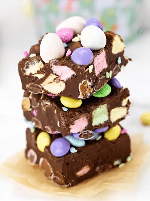 Stack of colorful no-bake Easter-themed dessert bars for a bake sale, made with chocolate fudge, marshmallows, candy-coated chocolates, and topped with pastel Easter eggs and sprinkles.