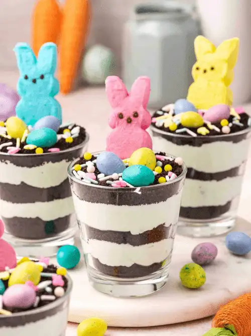 Layered no-bake Easter-themed desserts for a bake sale, featuring chocolate cookie crumbs and cream in small dessert cups topped with pastel candy eggs, sprinkles, and colorful marshmallow bunny candies.