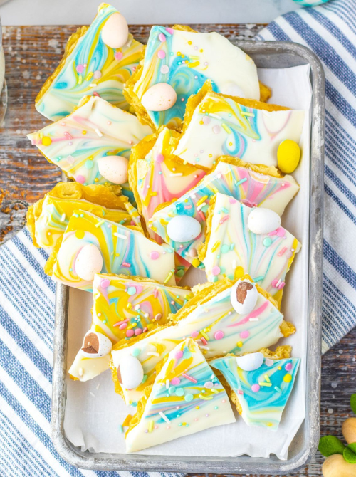 Tray of colorful no-bake Easter-themed dessert bark for a bake sale, featuring swirled white chocolate with pastel candy eggs, sprinkles, and bright spring colors broken into festive pieces.