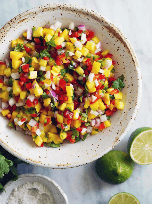 Easy Cinco de Mayo party food recipes for a crowd featuring a vibrant bowl of fresh mango salsa with diced mango, red onion, jalapeño, cilantro, and lime on a light marble surface.
