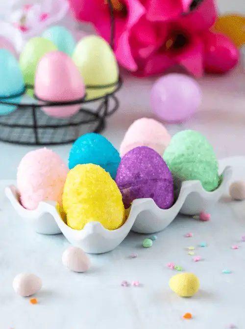 Colorful no-bake Easter-themed desserts for a bake sale, featuring sugar-coated pastel Easter egg treats arranged in a ceramic egg tray with festive spring decorations in the background.