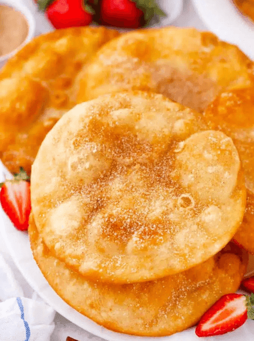 Golden crispy Mexican buñuelos dusted with cinnamon sugar stacked on a white plate with fresh strawberries — an easy Cinco de Mayo party food recipe for a crowd that makes 8 hand-held fried desserts for under $6.