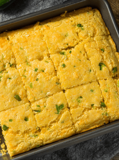 Jalapeño cheddar Mexican cornbread cut into 16 squares in a metal baking pan showing a golden, moist crumb studded with green jalapeño pieces — an easy Cinco de Mayo party food recipe for a crowd that bakes in just 25 minutes.
