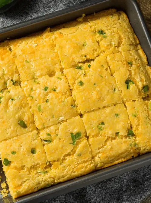 Jalapeño cheddar Mexican cornbread cut into 16 squares in a metal baking pan showing a golden, moist crumb studded with green jalapeño pieces — an easy Cinco de Mayo party food recipe for a crowd that bakes in just 25 minutes.