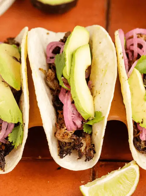 Instant Pot pork carnitas tacos in flour tortillas topped with crispy shredded pork, pickled red onions, fresh avocado slices, and cilantro — an easy Cinco de Mayo party food recipe for a crowd that serves 10 with just 5 minutes of prep.