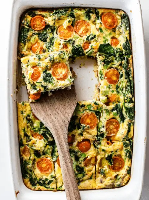 Spinach, tomato, and egg breakfast casserole baked in a white dish with a slice being served, a fresh veggie-packed option for make-ahead Easter brunch casseroles.