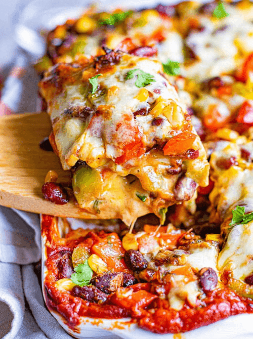 Vegetarian enchilada casserole being served with a wooden spatula showing layers of melted Monterey Jack cheese, red beans, corn, and colorful bell peppers in enchilada sauce — an easy Cinco de Mayo party food recipe for a crowd that's naturally vegetarian and make-ahead friendly.