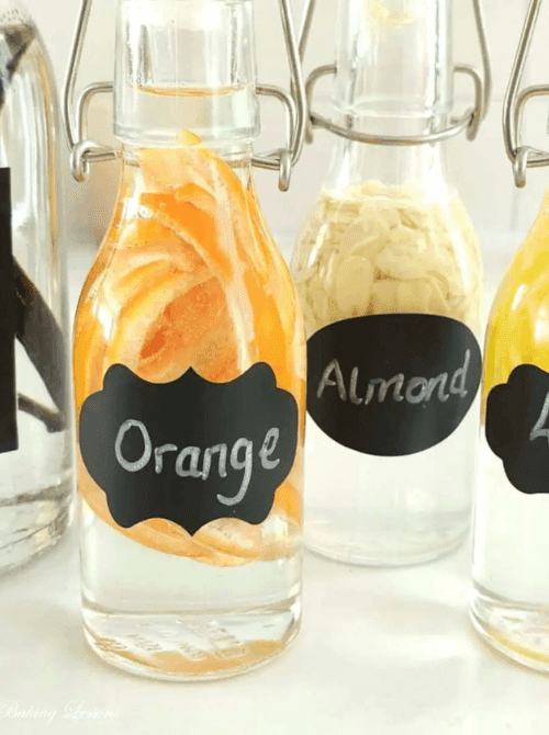 Homemade alcohol-free baking extracts in swing-top glass bottles with chalkboard labels showing orange peel and almond — a handmade kitchen gift idea for DIY Mother's Day gift baskets.