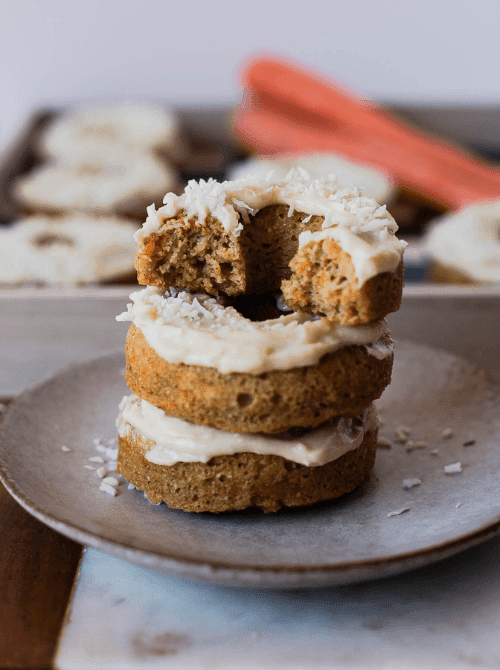 A stack of two baked carrot cake donuts topped with honey cream cheese frosting and shredded coconut on a rustic gray plate, with a bite taken from the top donut revealing a moist spiced crumb, fresh carrots and a donut pan visible in the background — a naturally gluten-free make-ahead Mother's Day brunch idea that preps in 15 minutes and satisfies guests with dietary restrictions without feeling like a compromise.