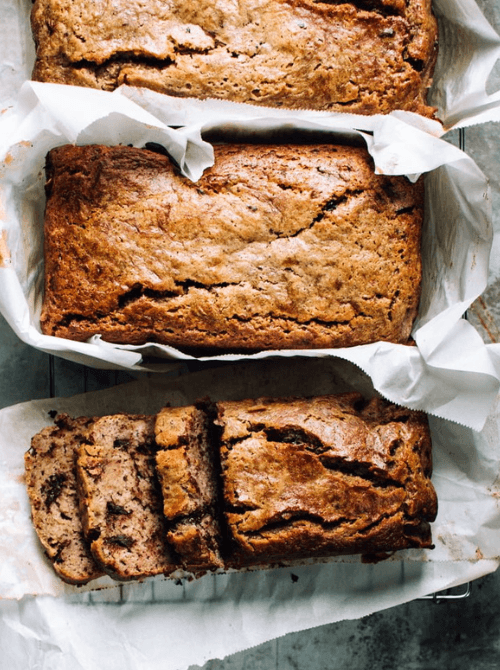 Two golden banana bread loaves in parchment-lined loaf pans from above, one whole and one partially sliced showing a moist tender crumb — a classic make-ahead Mother's Day brunch idea that stays soft for days, requires no mixer, and is ready to slice and serve straight from the pan.